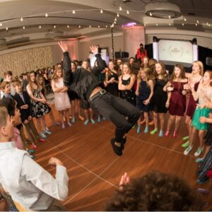 A man performing an energetic jump in the air during a lively BAR Mitzvah celebration, captivating a crowd of people.