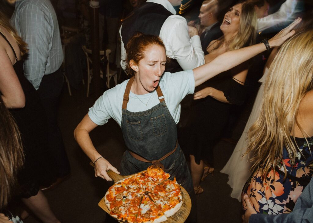 A woman in an apron holding a pizza at a party.