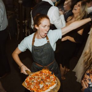 A woman in an apron holding a pizza at a party.