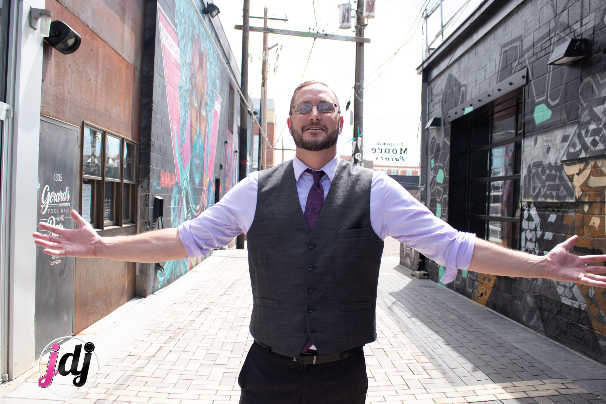 A man wearing a vest and tie, representing a DJ service, standing in an alley.