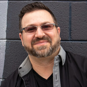 A man with glasses and a beard smiling in front of a wall at a bar mitzvah.