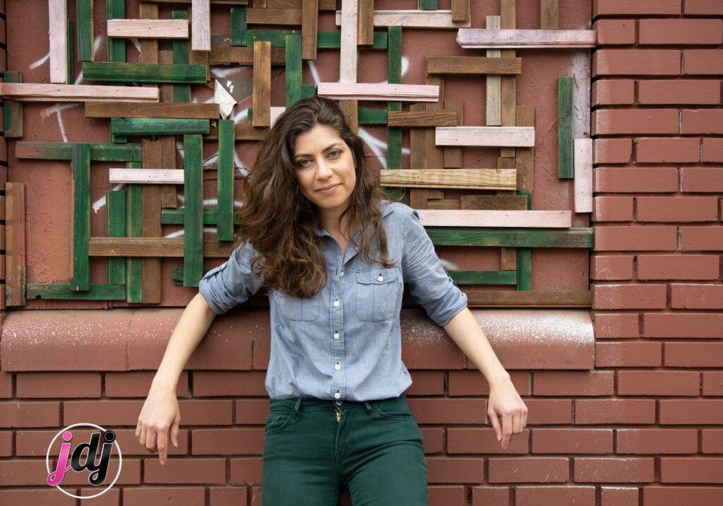 A woman in green pants leaning against a brick wall at a bat mitzvah.