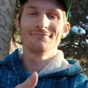 A young man in a plaid shirt and cap, giving a thumbs up to the camera with trees and a brick building in the background, potentially planning event entertainment.