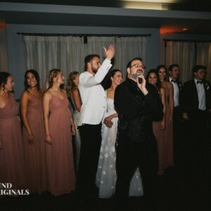 A wedding party stands in a line indoors, captured beautifully for the wedding gallery. A man with a microphone is speaking while the bride and groom wave in the background. Bridesmaids are in matching dresses, and groomsmen are in suits.