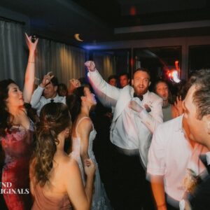 In a dimly lit room akin to an art gallery, a group of people are dancing energetically. Some are raising their hands while others are smiling and enjoying the moment, reminiscent of a joyous wedding celebration.
