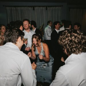 A group of people, mostly in white shirts and formal wear, are dancing in a dimly lit room with curtained windows. The atmosphere feels lively and energetic, reminiscent of a wedding photo gallery capturing joyous moments.