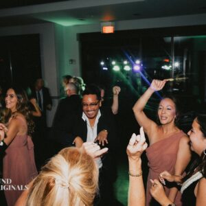 A group of people, dressed in formal attire, are dancing and celebrating at an indoor event. The room is dimly lit like an elegant gallery, with colorful lights and decorations in the background, adding to the festive wedding atmosphere.