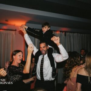A man with a child on his shoulders dances with others in a lively indoor setting, featuring dim lighting and semi-formal attire, reminiscent of a vibrant wedding reception.