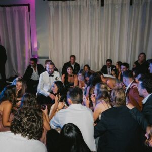 A group of people, dressed in formal attire, are sitting closely together on the floor at an indoor wedding event with curtains and purple lighting in the background.