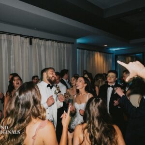 A group of people, including a bride and groom, are seen dancing and celebrating in a dimly lit indoor wedding setting. The groom is wearing a white tuxedo, and the bride is in a white dress—a true gallery of love and joy.