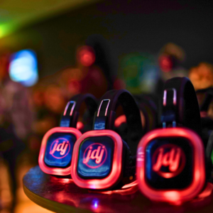 Several LED-lit headphones rest on a table, ready to transform the space into a vibrant silent disco, with a blurred background of people and ambient lighting setting the scene.