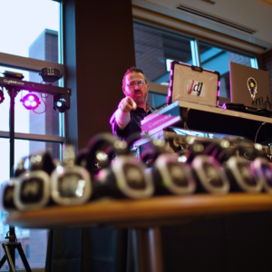 DJ at a booth, pointing towards the camera, with a row of wireless headphones on a table in the foreground, setting the stage for an unforgettable silent disco.