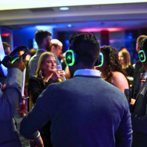 In a dimly lit room, people wearing glowing headphones immerse themselves in lively conversations and sip drinks, creating the unique ambiance of a silent disco.