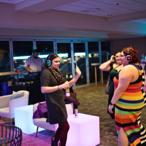 Guests in vibrant attire enjoy a unique silent disco with headphones in a lounge setting, while a DJ crafts the beats in the background.