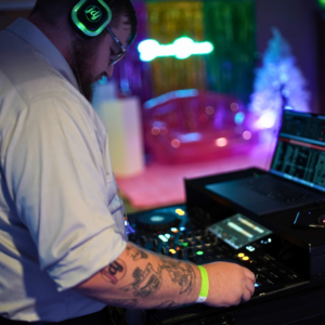 A DJ with headphones operates a mixer at a vibrant event, reminiscent of a lively silent disco, with colorful lights and decorations in the background.