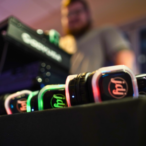 Several LED-lit wireless headphones are lined up on a table, creating a perfect setup for a silent disco, with a DJ standing in the background near sound equipment.