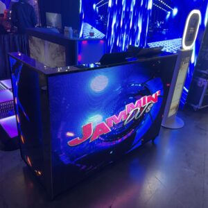 A DJ booth with a lit-up display showing "Jammin DJs" in neon colors, set in a dimly lit event space with blue lights.