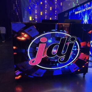 Large illuminated DJ booth with a colorful digital display showing the letters "jdj" in a circular design. Bright stage lights are visible in the background.