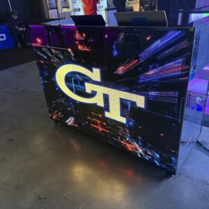 Illuminated display with the letters "GT" on a digital backdrop, placed on a concrete floor, with a booth and colorful lights in the background.