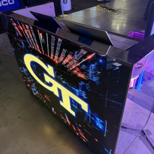 A DJ booth with a large screen displaying "GT" in bright colors. Two laptops are on top of the booth in an indoor event setting.