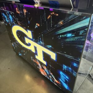 Large screen displaying "GT" with a digital cityscape background, positioned on a table.