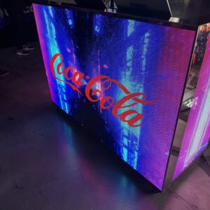An illuminated counter displays the Coca-Cola logo in a vibrant, digital setting with a purple and blue background.