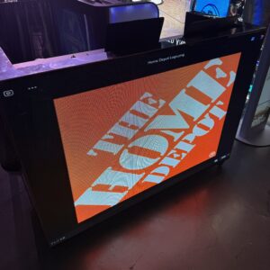 A large electronic display with "The Home Depot" logo is illuminated in an event space with a basketball game in the background.