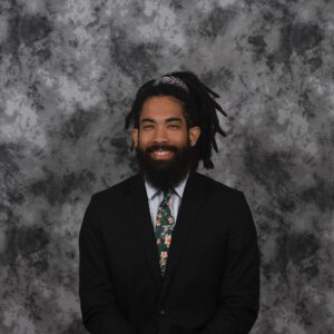 A man in a suit with a floral tie smiles against a gray marbled backdrop. He has a beard, his hair in locks tied back, reminiscent of professional DJs who effortlessly blend style and charisma.