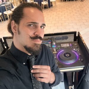 A man with a mustache and beard stands beside DJ equipment in an event space. He holds his tie with one hand. The room has tables with black tablecloths and gold chairs.