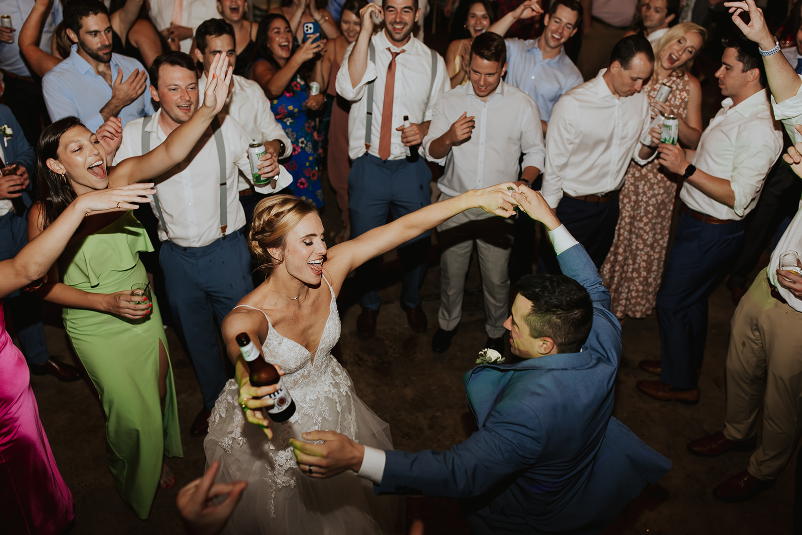 A bride and groom dance surrounded by guests at a lively wedding reception, with people smiling, cheering, and holding drinks.