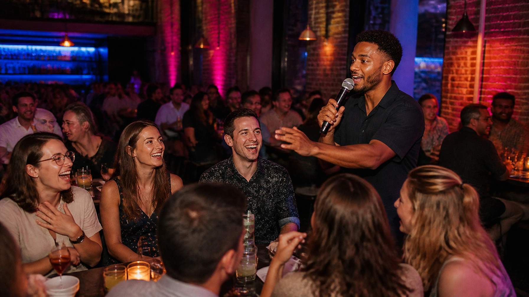 A comedian performs with a microphone for a laughing audience at a crowded, dimly lit comedy club with exposed brick walls and colorful lighting.