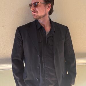 A man wearing sunglasses and a black blazer stands against a beige wall, looking to the side with his hands in his pockets.