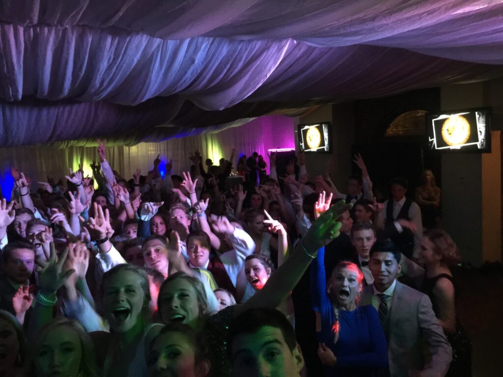 A large group of people dressed formally are dancing and raising their hands at an indoor event with colorful lighting and draped ceiling fabric.