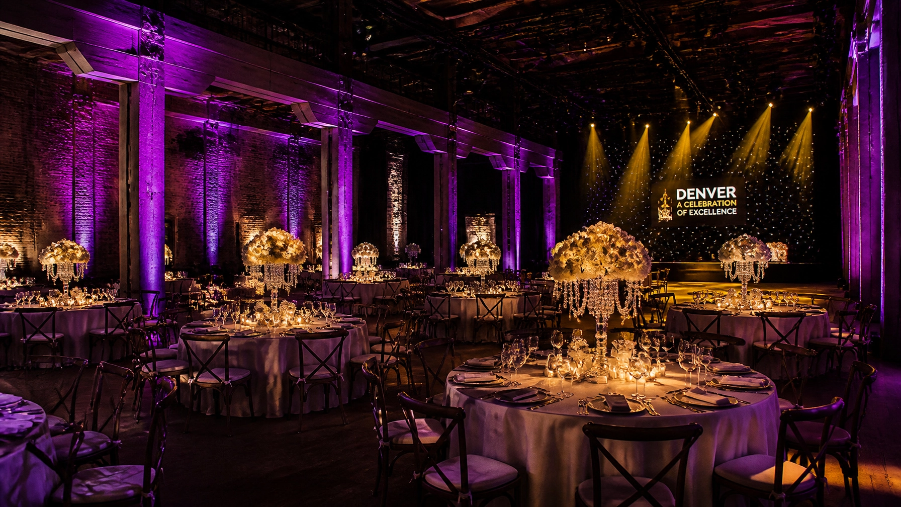 Elegant banquet hall with round tables, crystal centerpieces, and dramatic purple lighting. A stage with “Denver Celebration of Excellence” is visible in the background.
